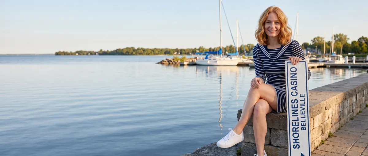 Generated image: the guide's character, a young woman with strawberry-blonde hair in a navy-and-white striped sailor mini dress, sits on a stone wall by a marina on a calm bay at golden hour, holding a white sign that reads Shorelines Casino Belleville.